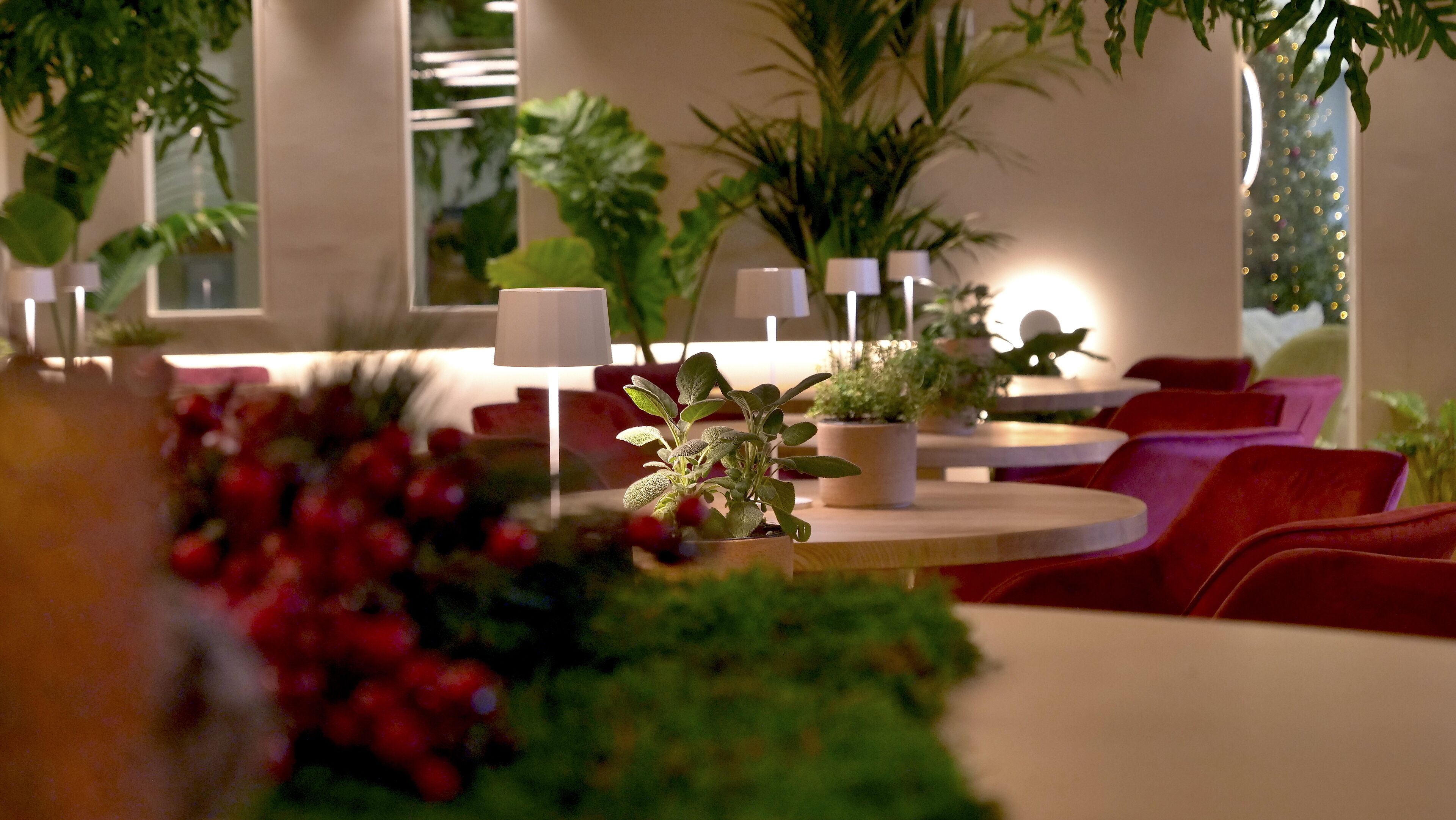 A modern indoor setting with round tables, red upholstered chairs, and potted plants.