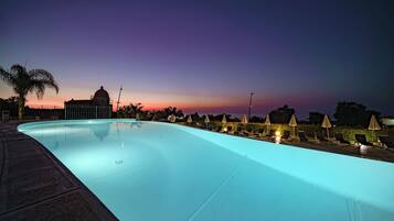 Una piscina al anochecer con sillas de descanso y sombrillas, una palmera y un edificio de forma abovedada al fondo.