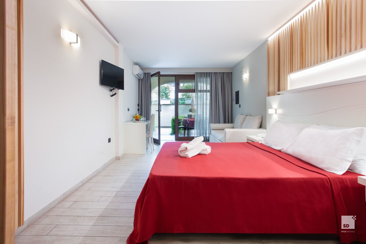 A hotel room with a red bedspread, a flat-screen TV mounted on the wall, and a view of the outdoor area through a glass door.