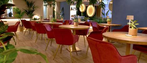 A restaurant interior with round tables and red chairs, featuring potted plants and a bar area.