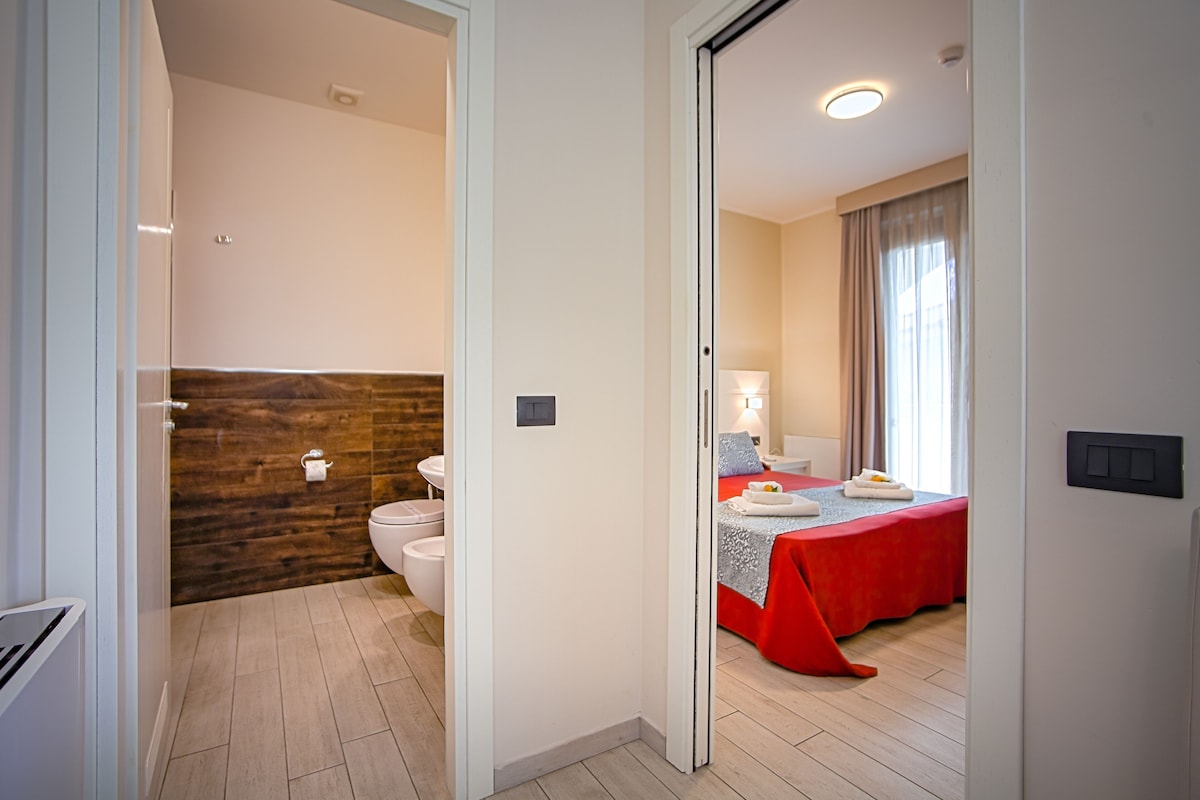 Une chambre d’hôtel avec un mur en bois, un lit recouvert d’une courtepointe rouge, une salle de bain avec des toilettes et un lavabo, et une fenêtre avec des rideaux.