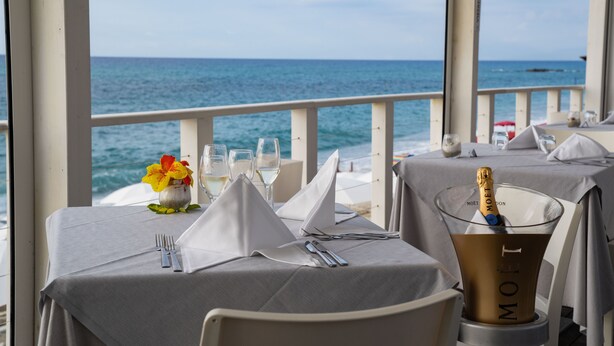 Une table dressée pour le dîner, avec vue sur l’océan, une bouteille de champagne et un arrangement floral en guise de centre de table.