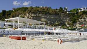 Une plage avec des parasols blancs et des chaises longues, un panneau indiquant « Plage Blanca », et une falaise rocheuse en arrière-plan.