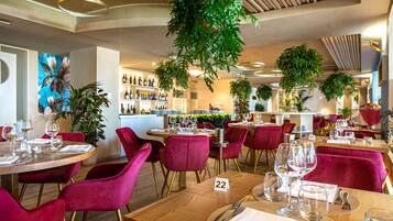 A restaurant interior with round tables, red velvet chairs, and a variety of plants.