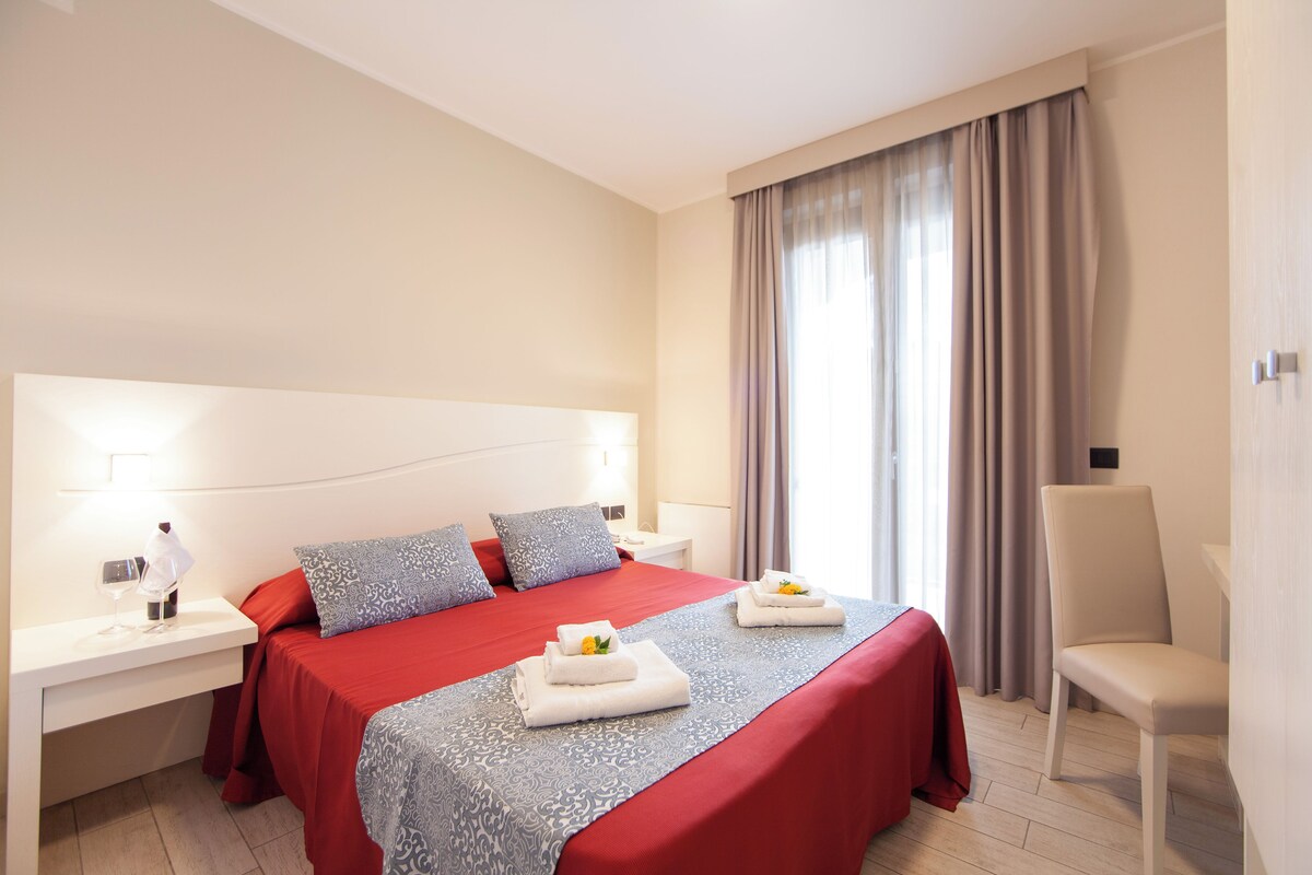 A hotel room with a large bed, a bedside table with a lamp, a chair, and a window with curtains.
