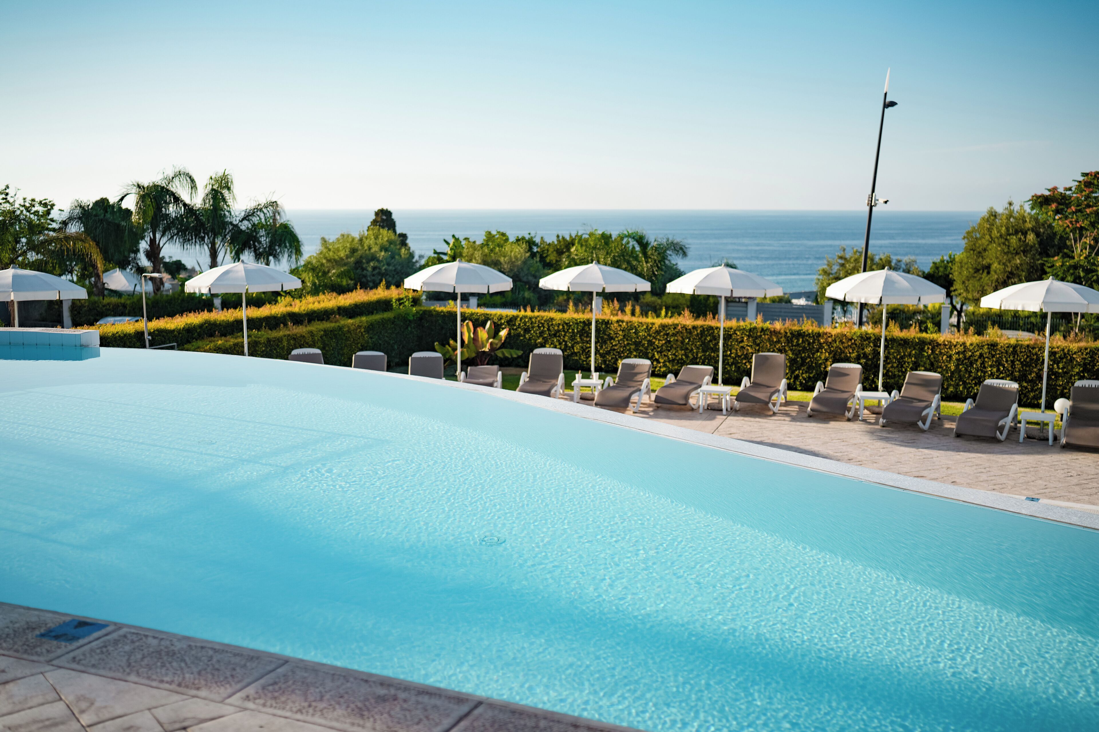 A pool area with lounge chairs and umbrellas, a clear view of the ocean, and well-maintained landscaping.