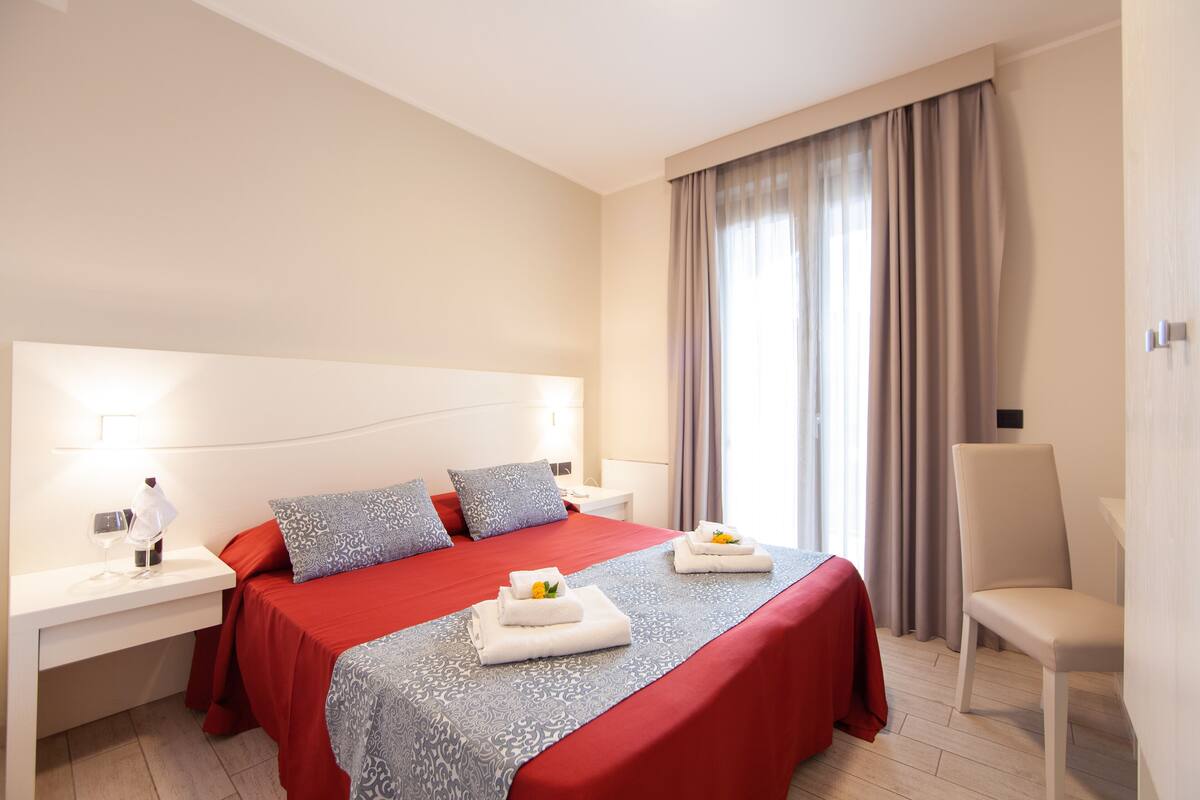 Une chambre d’hôtel avec un grand lit, une table de chevet avec une lampe, une chaise et une fenêtre avec des rideaux.
