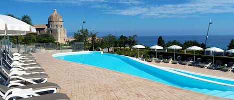 A swimming pool with lounge chairs and umbrellas, a stone pathway, and a view of the sea and a historic building in the background.