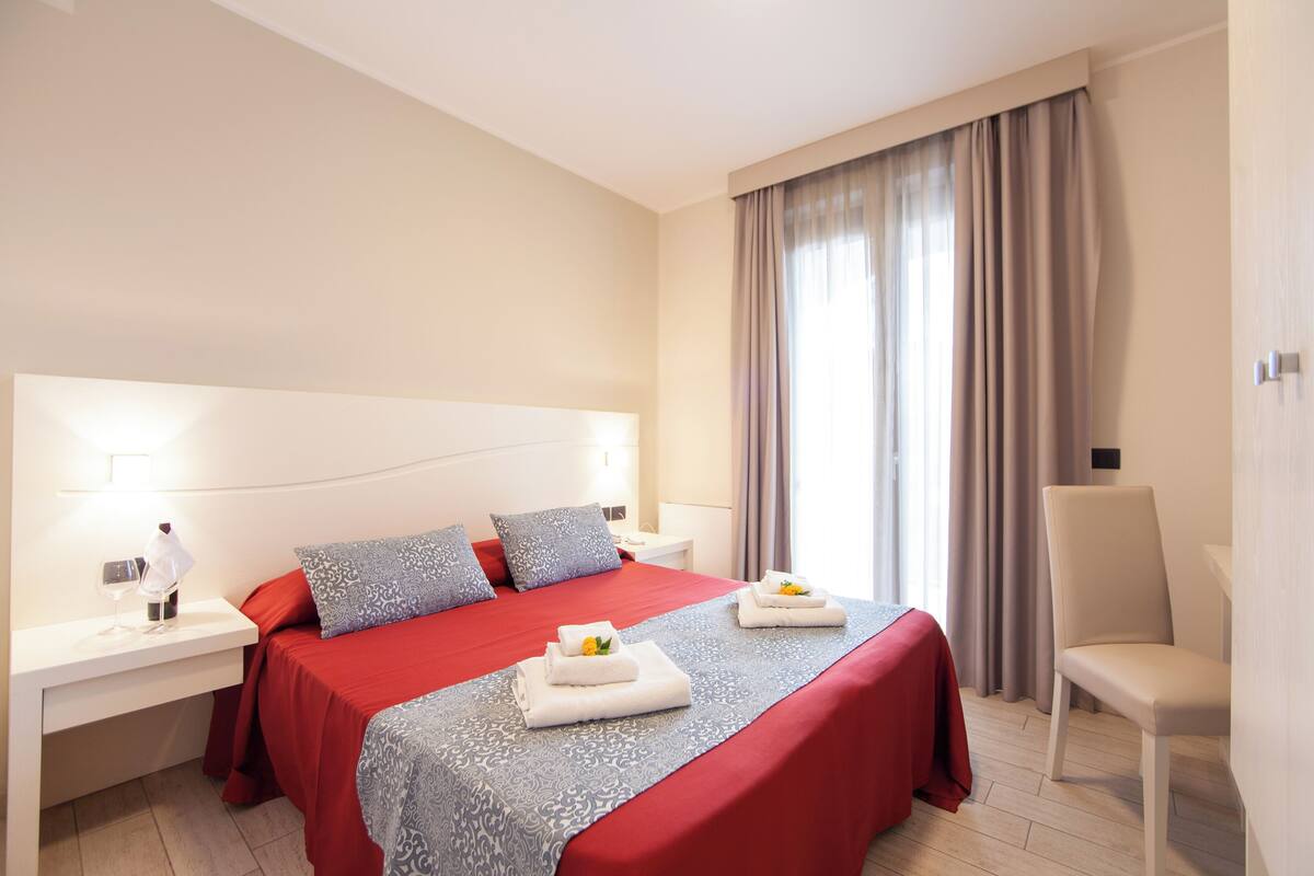Une chambre d’hôtel avec un grand lit, une table de chevet avec une lampe, une chaise et une fenêtre avec des rideaux.
