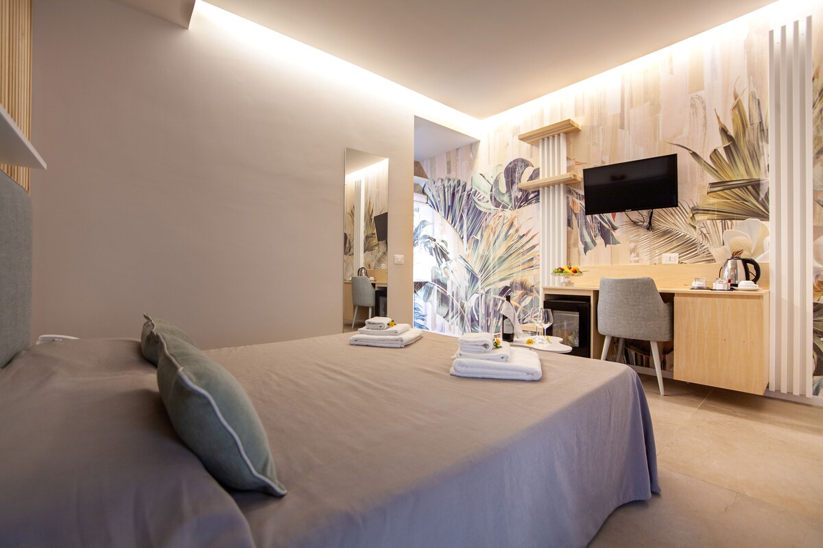 A hotel room with a bed, a desk, a chair, a TV, and a wall with a tropical wallpaper.