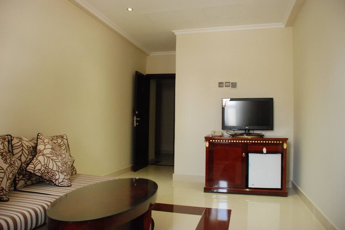 A living room with a sofa, a coffee table, a television on a wooden cabinet, and a door leading to another room.