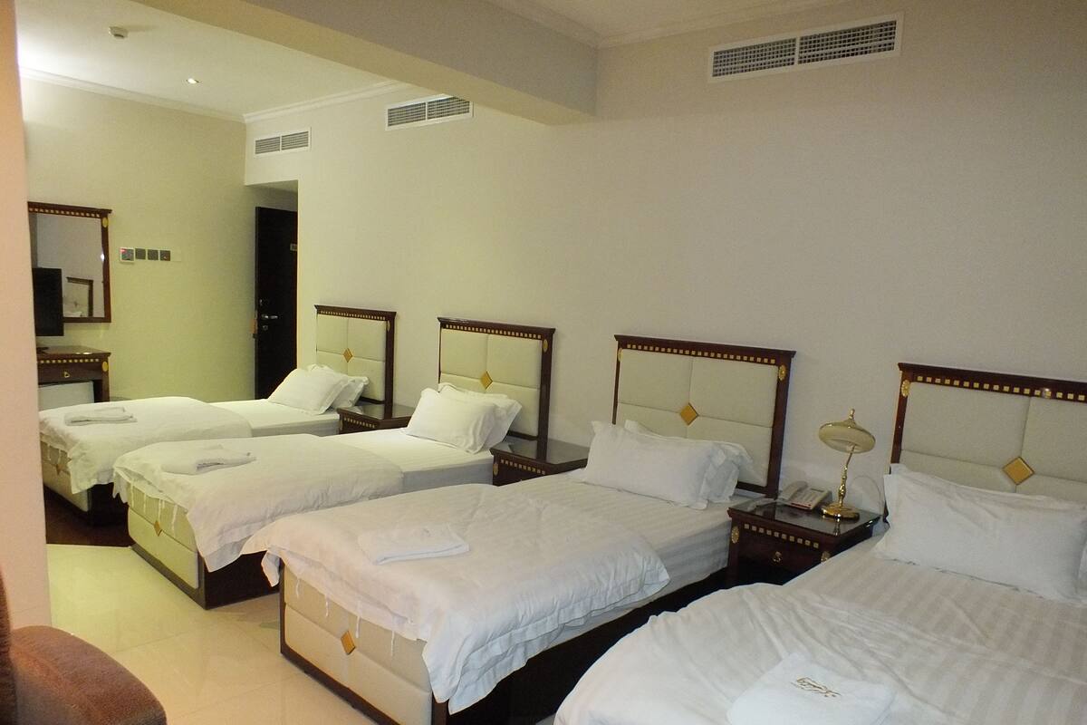 A hotel room with four single beds, each with a wooden headboard and white bedding. There are bedside tables with lamps, a mirror, and a small clock on the wall.