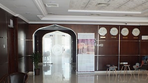 A hotel lobby with a reception desk, a large glass door entrance, a plant, and clocks displaying different time zones. - City Hotel Salalah (Salalah)