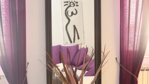 Une pièce dans laquelle on trouve, au mur, une œuvre d’art abstraite encadrée, une table en verre ornée d’un vase contenant des branches décoratives, et des rideaux violets.