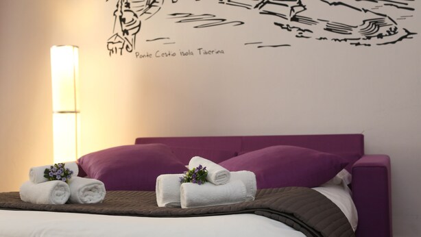 Une chambre d’hôtel avec un lit, une literie violette, une fresque murale et une lampe.