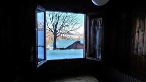 Ein Zimmer mit großem Fenster, Holzwänden und Blick auf eine verschneite Landschaft mit Baum und Gebäude.