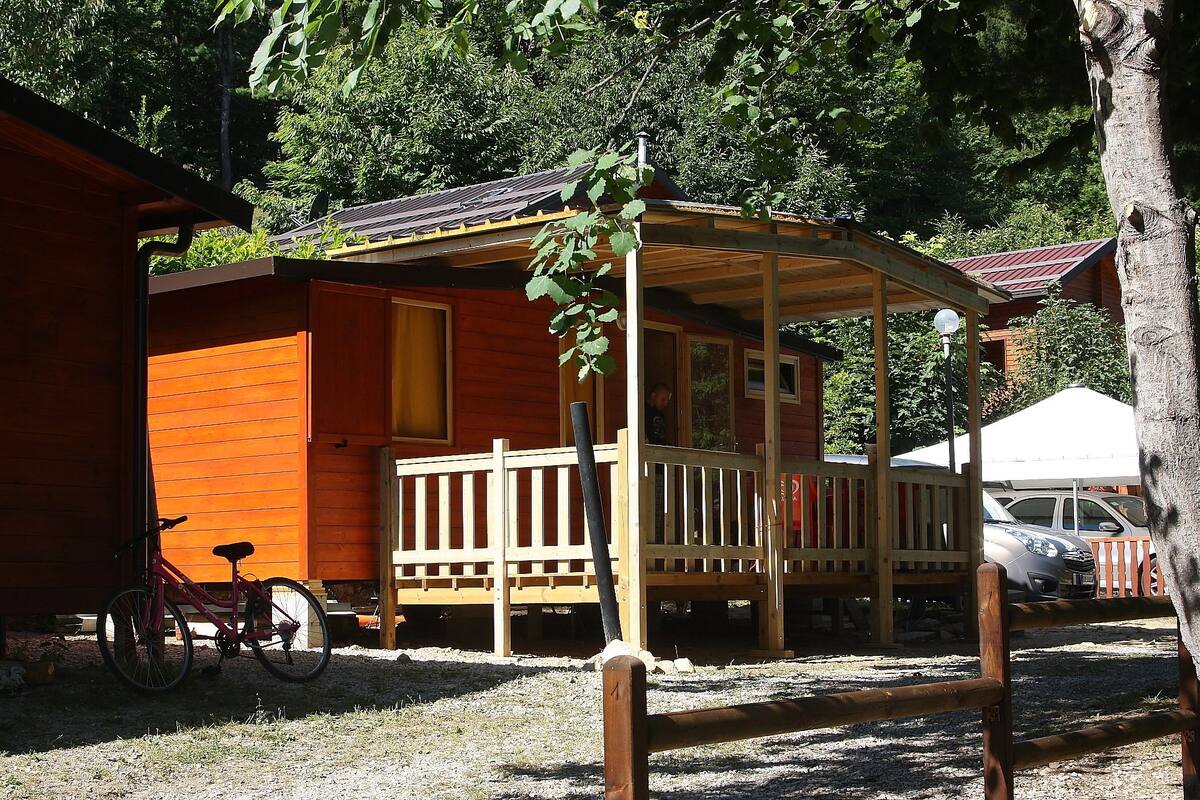 Una cabaña de madera con un porche, una bicicleta y un coche aparcado cerca.