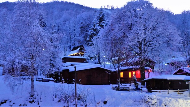 Ein mit Schnee bedecktes Chalet mit Holzfassade und beleuchtetem Dach, umgeben von schneebedeckten Bäumen und einem Bergpanorama.