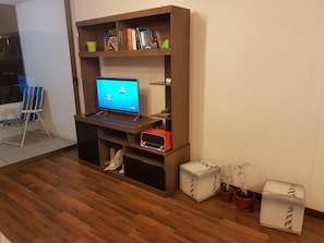 Quarto com piso de madeira, uma escrivaninha com computador, estante e uma cadeira.