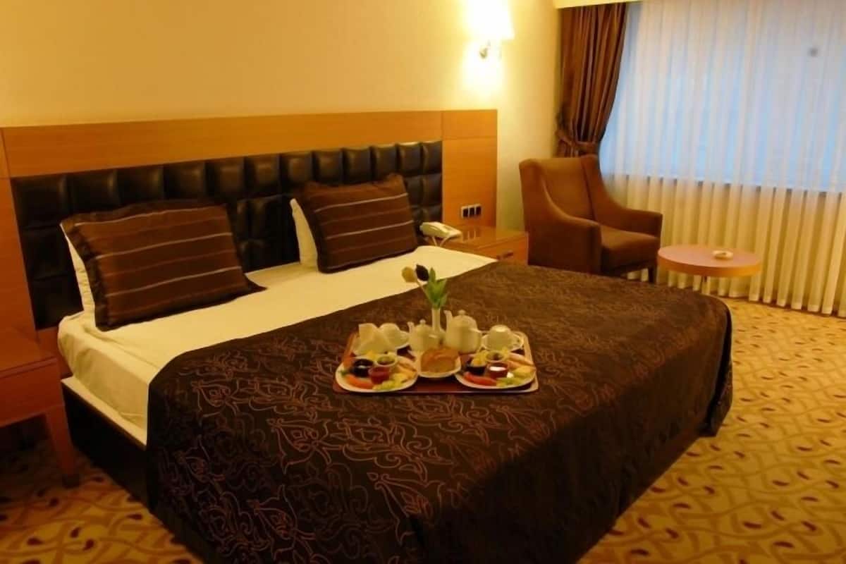 A hotel room with a large bed, a headboard, a bedside table with a phone, a chair, a small round table, and a tray with tea and snacks.