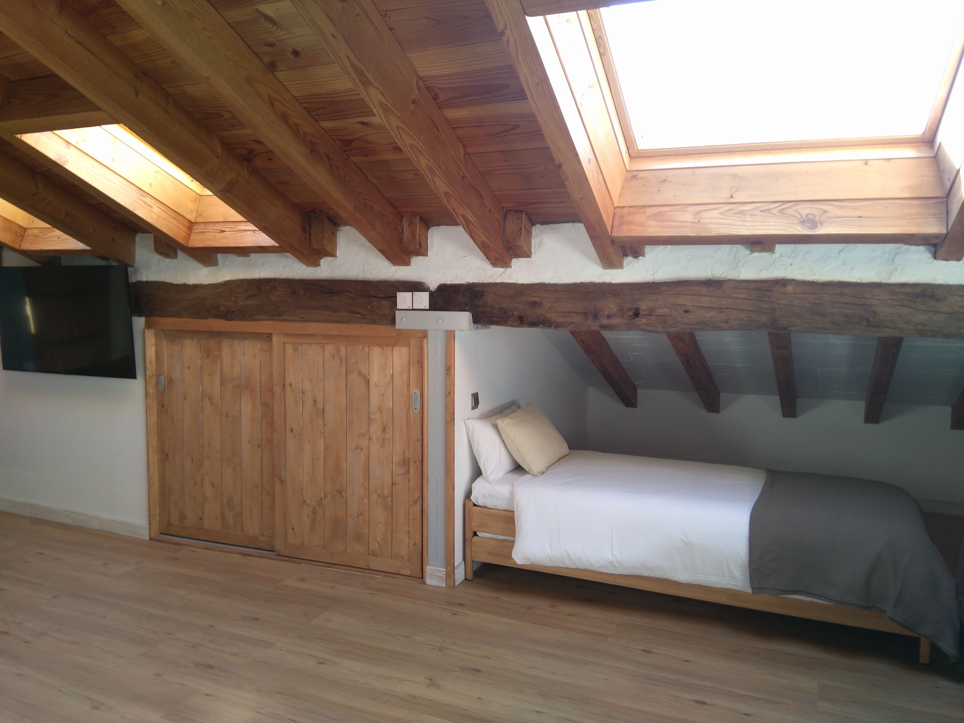 Une pièce avec un plafond en pente, des poutres en bois, un lit avec une literie blanche, une armoire en bois et un téléviseur fixé au mur.