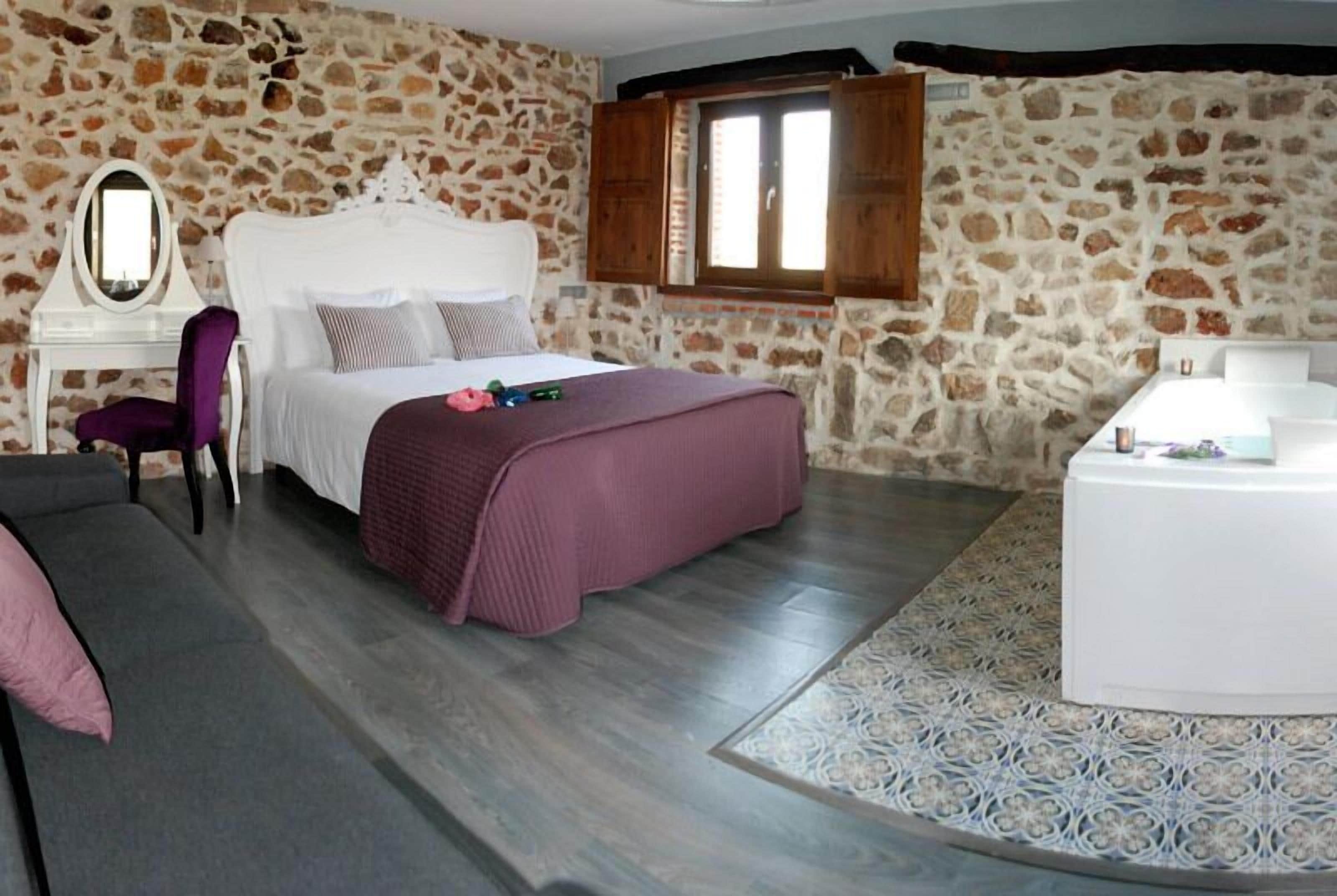 A bedroom with a stone wall, a bed with a white headboard, a purple chair, a small table with a lamp, and a decorative rug.