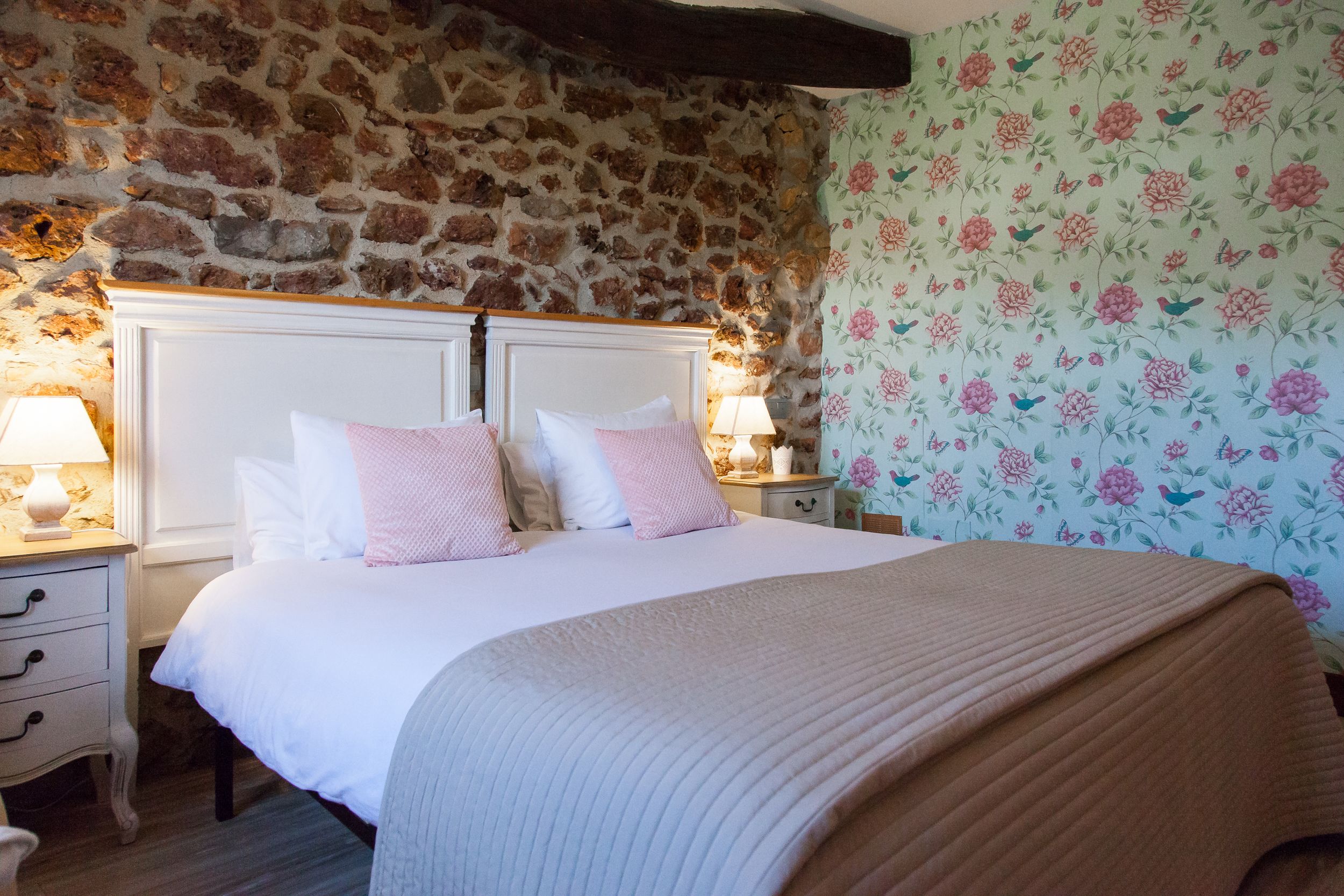 Ein Schlafzimmer mit Steinsandsteinwand, blumigem Wandpapier, einem weißen Bett mit rosa-weißer Bettwäsche und einem Holzboden.