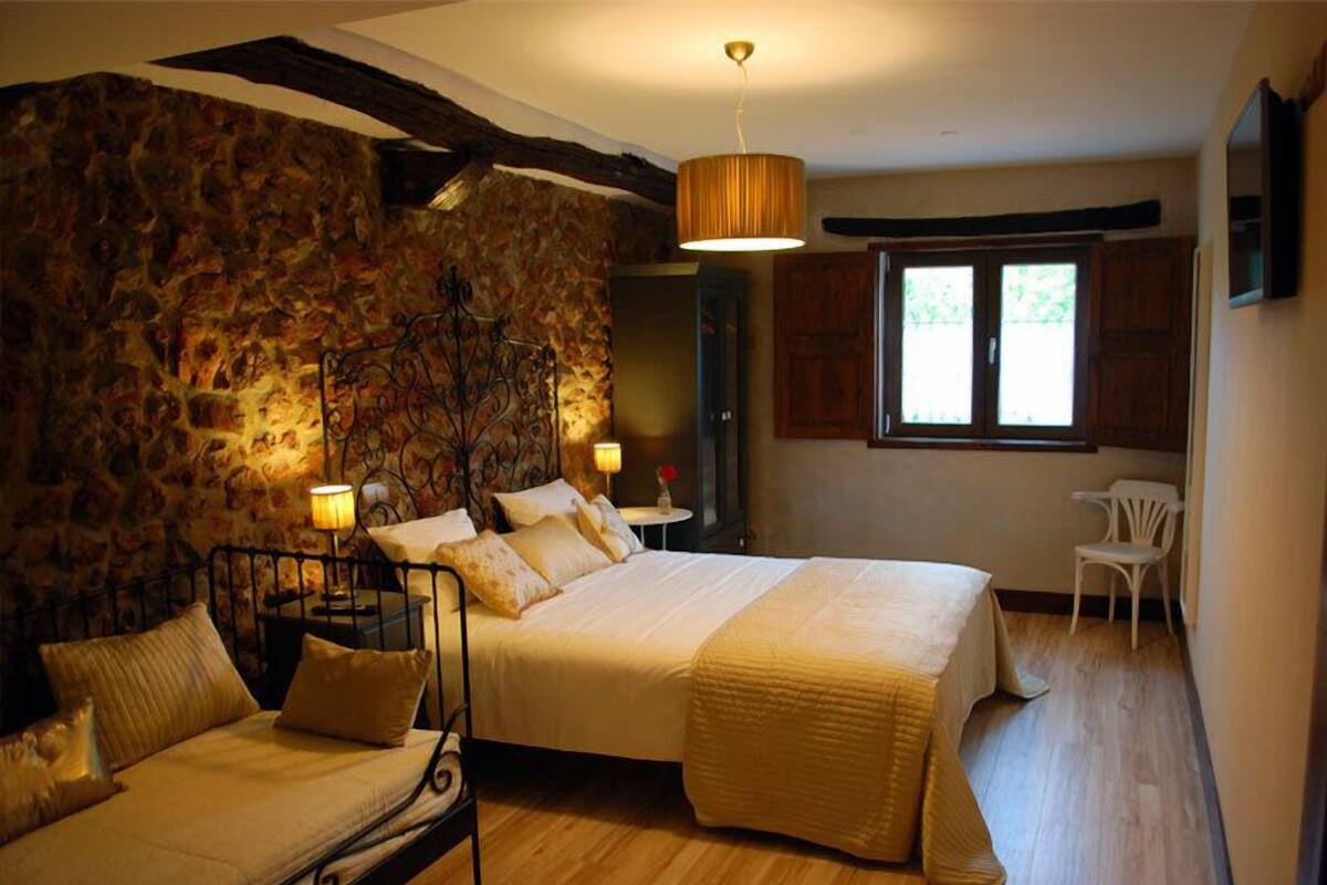 A bedroom with a stone wall, a bed with a canopy, a small table with a lamp, and a window with wooden shutters.
