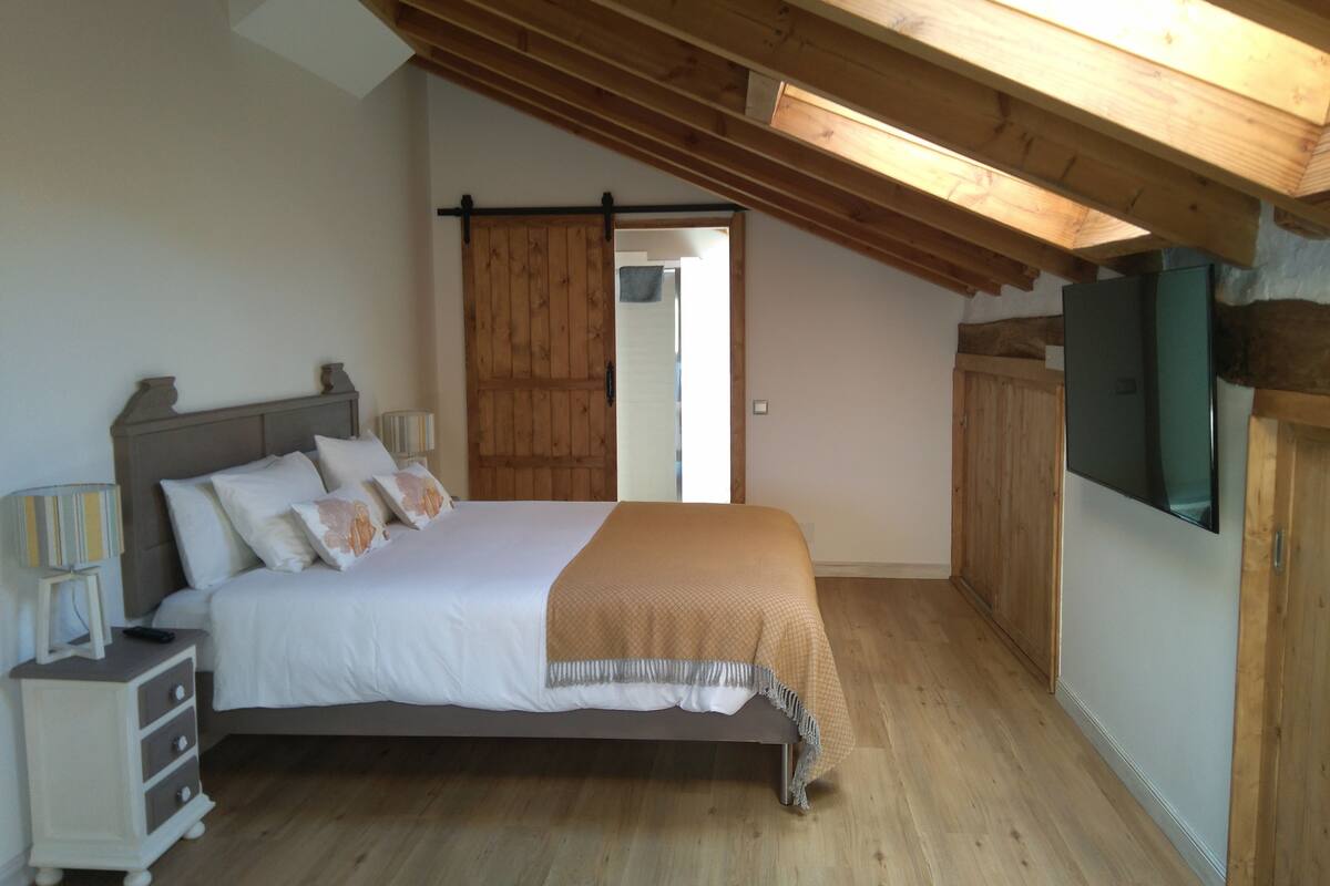 A bedroom with a wooden ceiling, a bed with white bedding, a nightstand with a lamp, and a wardrobe.
