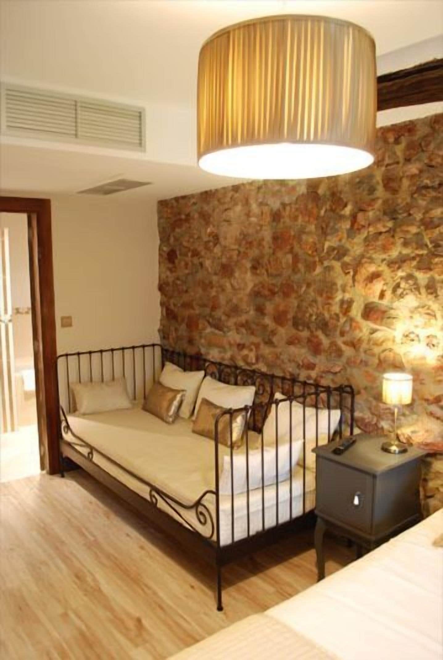 Un dormitorio con pared de piedra, cama con estructura de metal, mesita de noche con lámpara y lámpara en el techo.