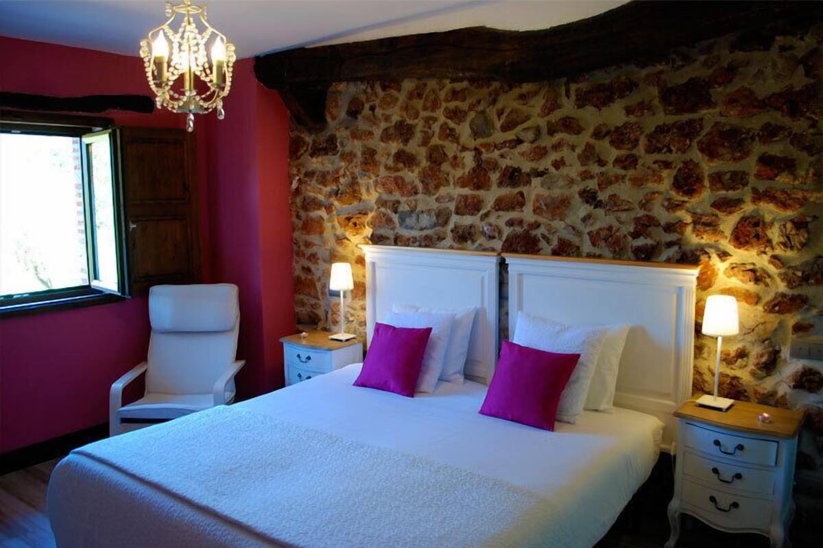 A bedroom with a stone wall, a white bed with purple pillows, a white chair, and a chandelier.