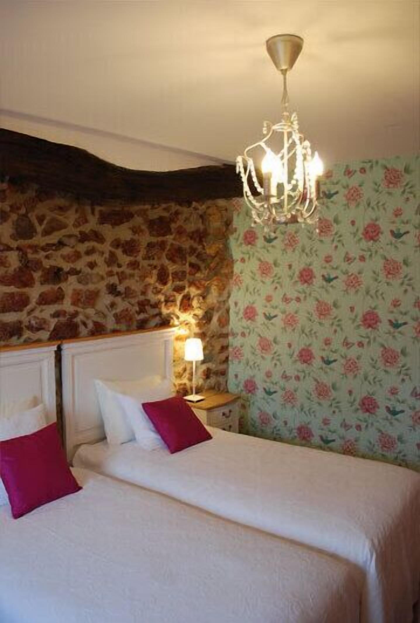 Un dormitorio con pared de piedra, papel tapiz floral, una araña y una cama con ropa de cama blanca y almohadas rojas.
