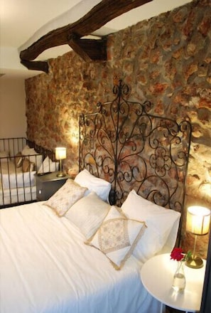 Quarto com parede de pedra, estrutura de cama em ferro forjado e mesa de cabeceira com abajur e um vaso de flores.