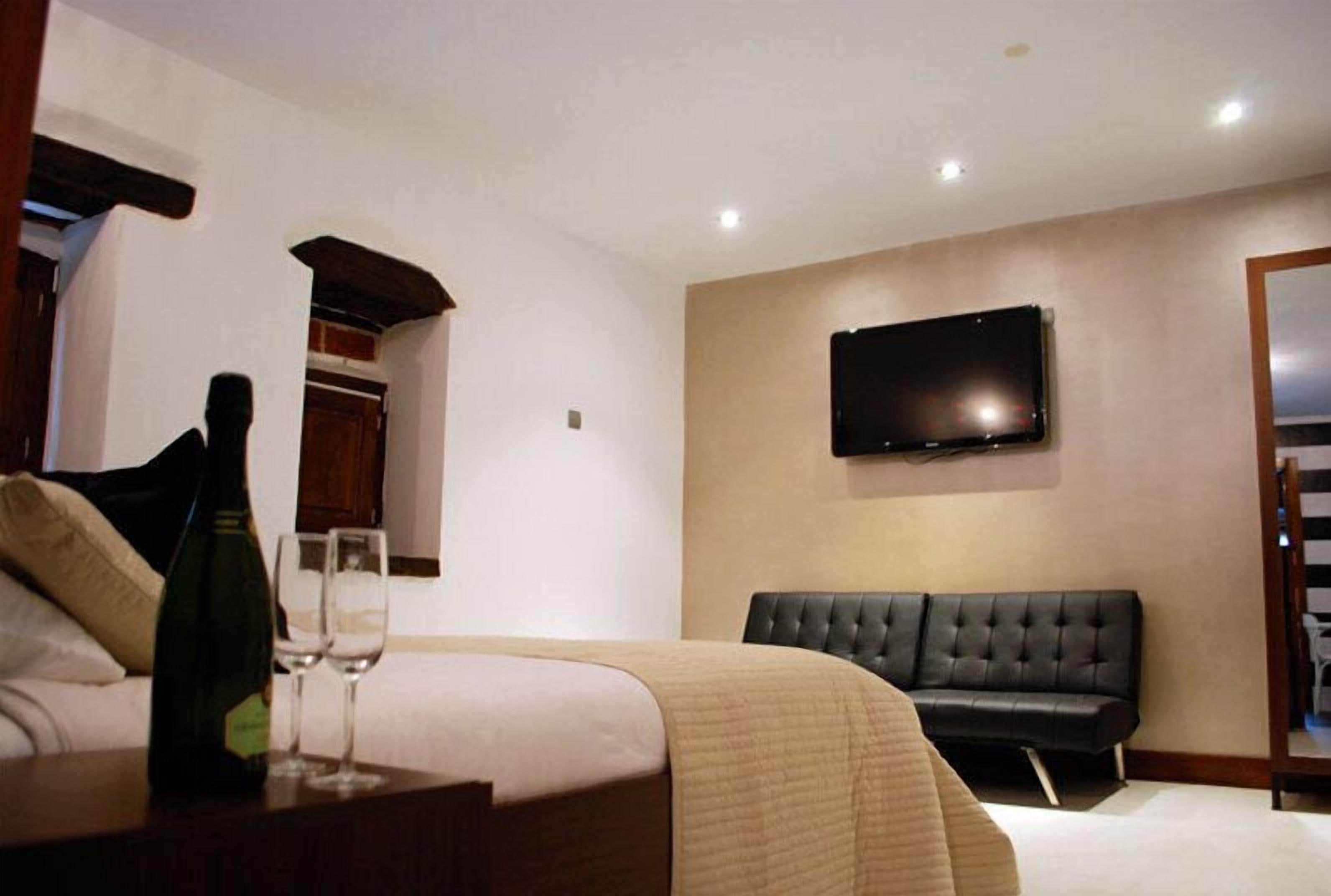 Habitación de hotel con cama, televisión montada en la pared, sofá de cuero negro y una botella de champán sobre la mesita de noche.