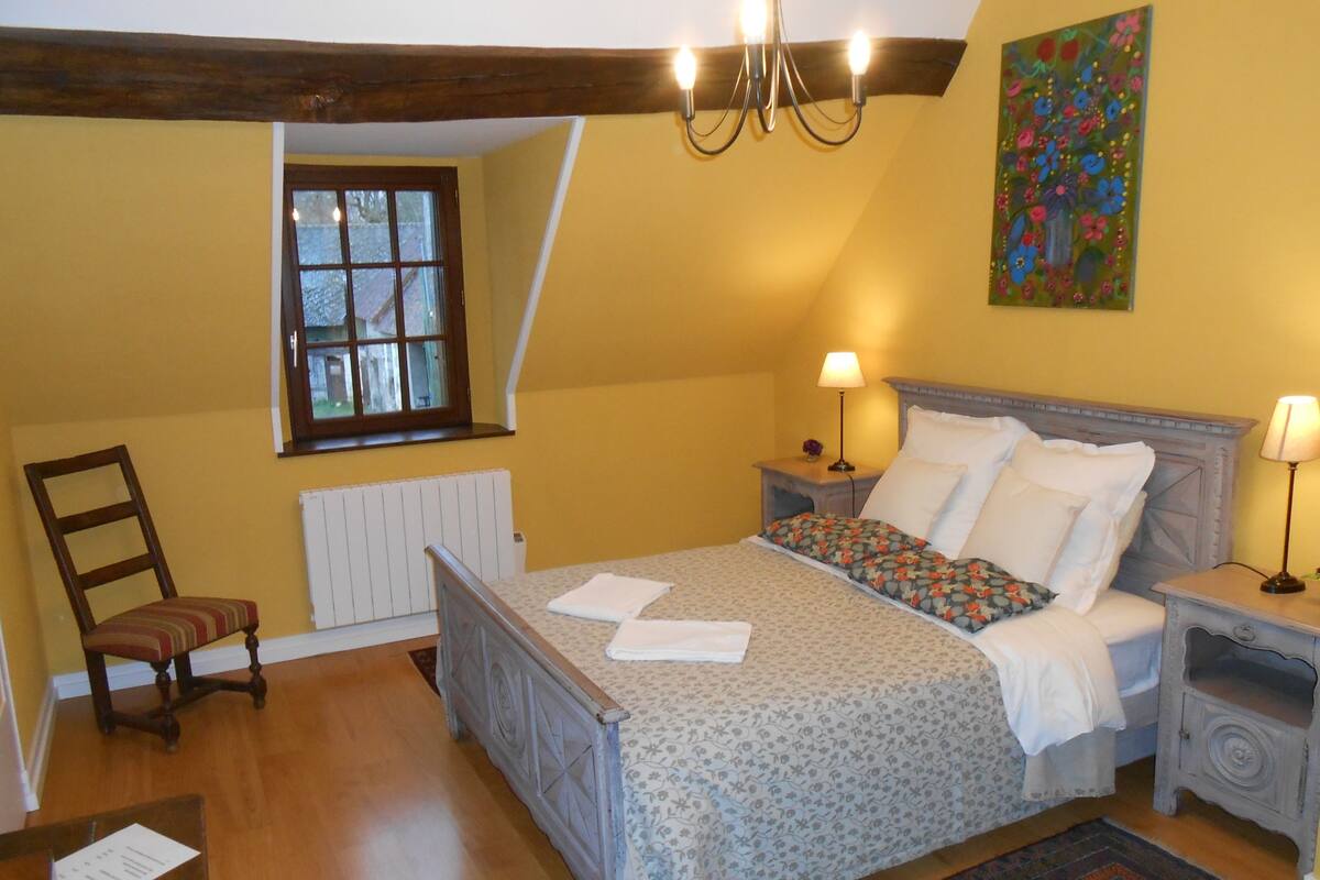 Un dormitorio con cama, dos mesitas de noche, una silla, un radiador, una ventana y una lámpara de araña.