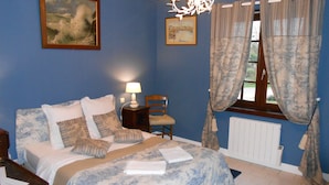 A bedroom with a bed, a chair, a nightstand, a lamp, a window with curtains, and a radiator. - B&B La Ferme Des Bordes (Pontlevoy)