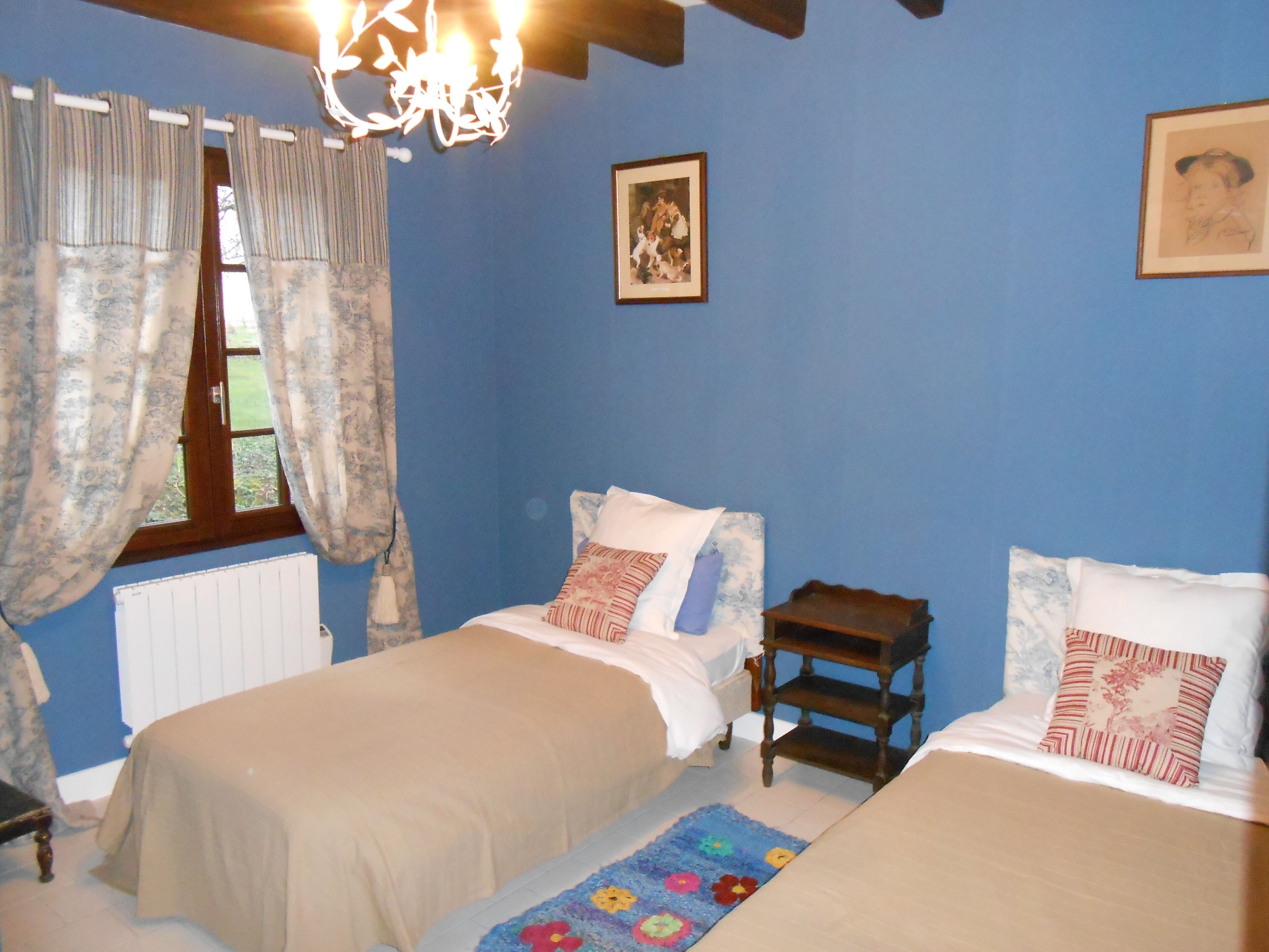 A bedroom with two beds, a window with curtains, a radiator, and a chandelier.