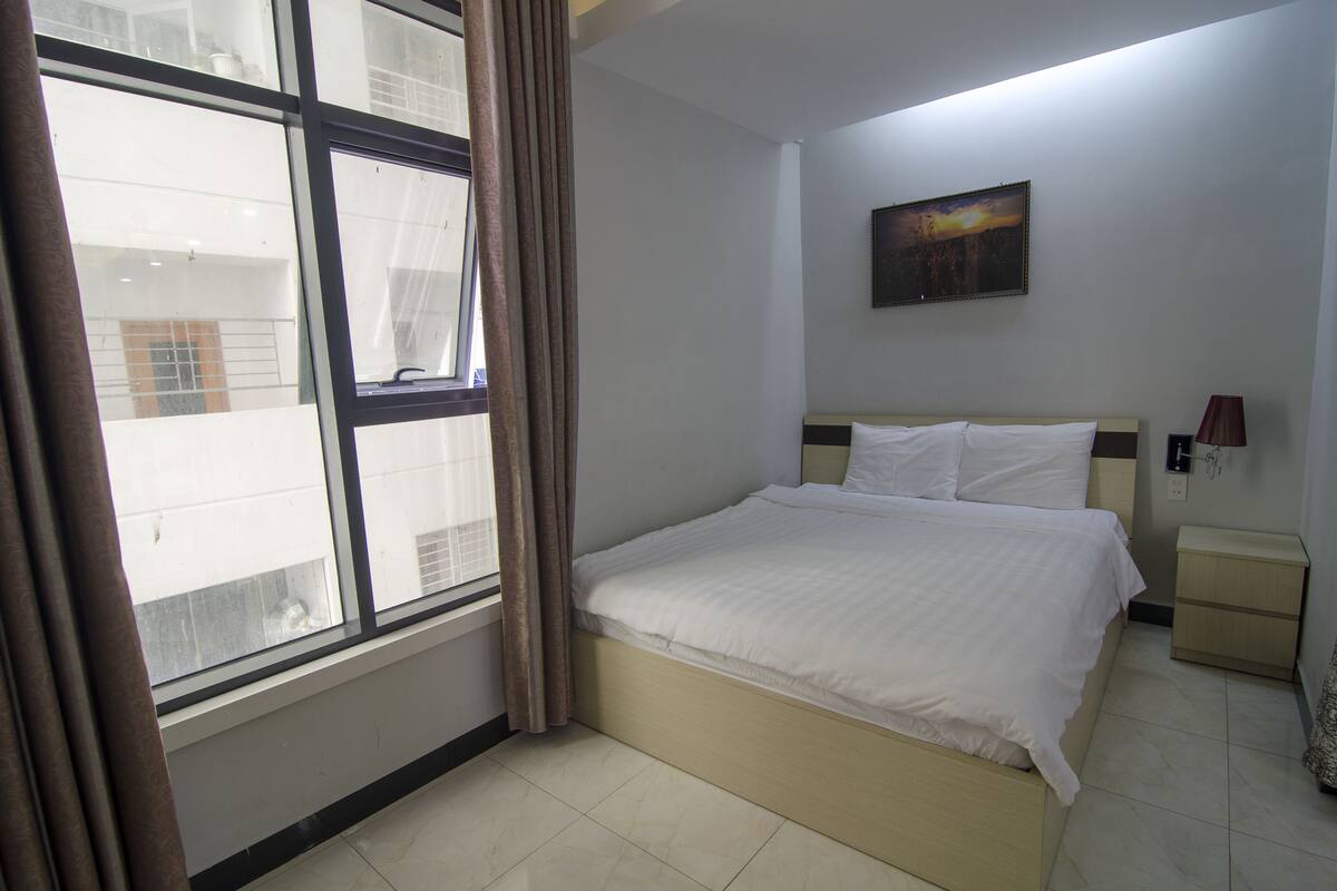 A bedroom with a bed, a nightstand, a lamp, a painting on the wall, and a window with curtains.