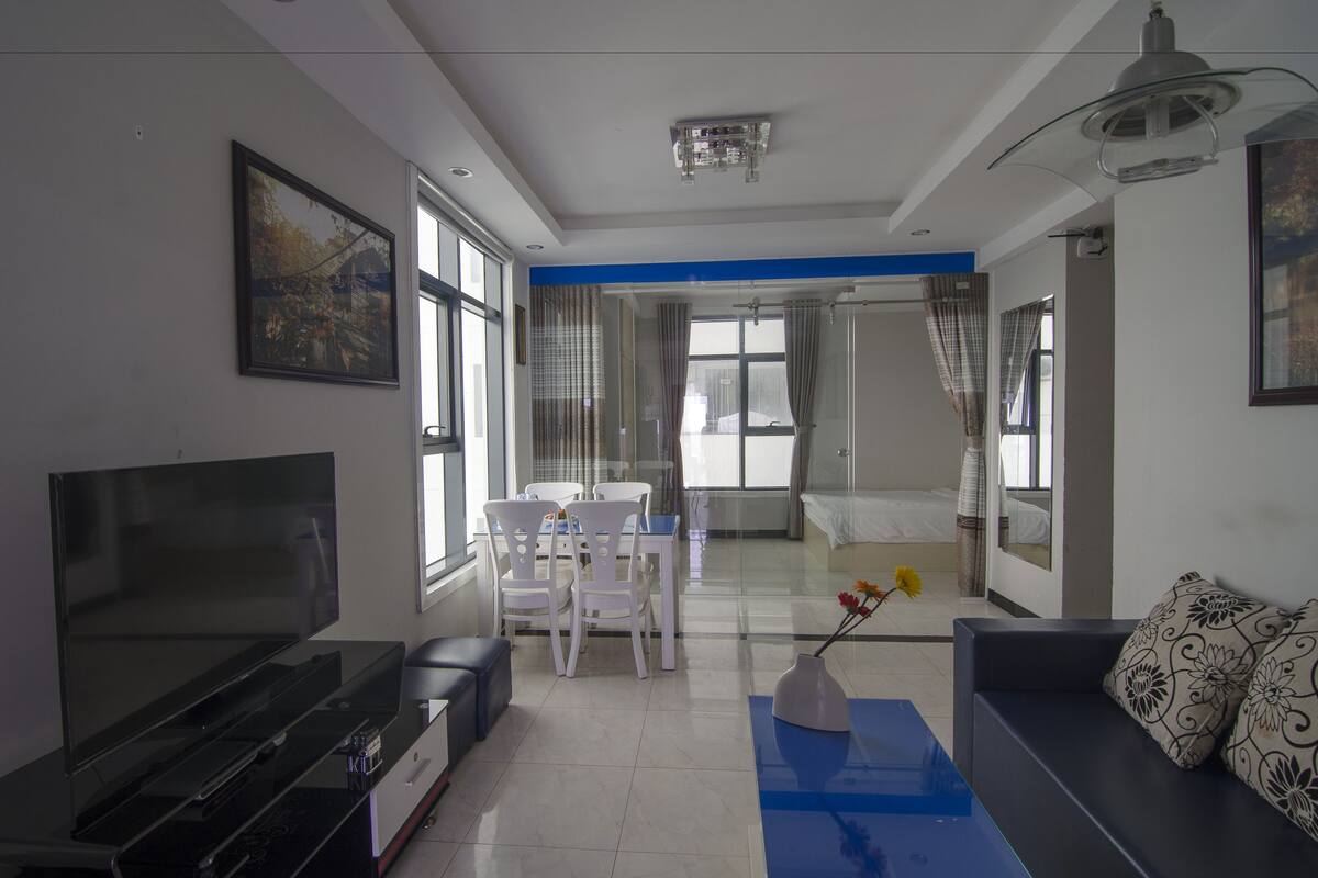 A modern living room with a flat-screen TV, a sofa, a dining table with chairs, and a large window with curtains.