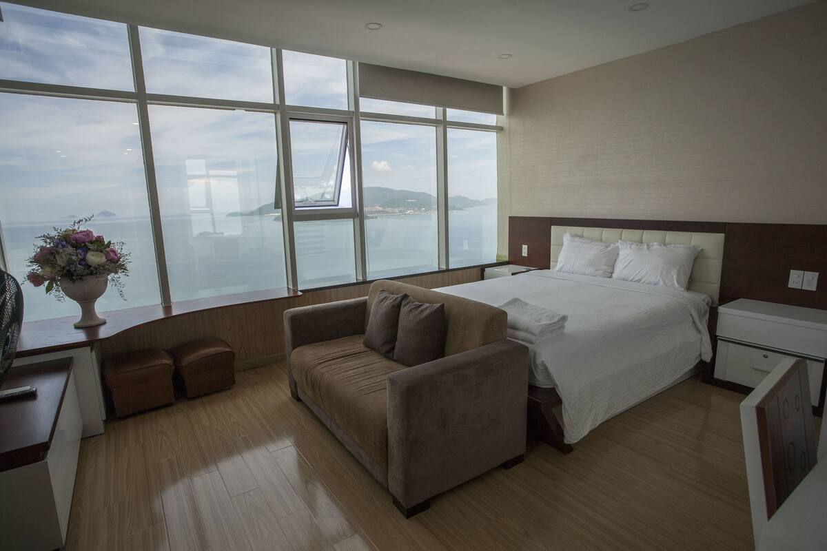A hotel room with a large window, a bed, a sofa, a nightstand, and a bedside table.