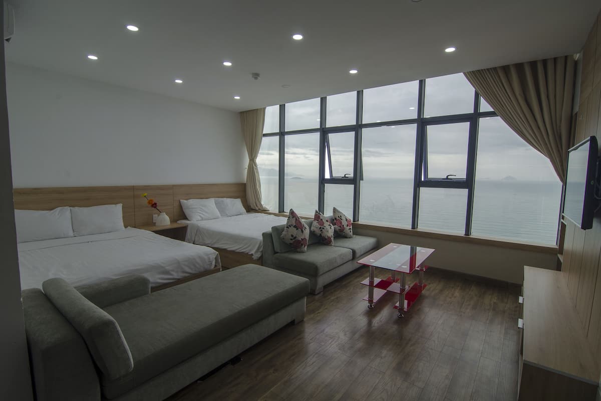 A hotel room with two beds, a sofa, a small table, and a large window offering a view of the sea.