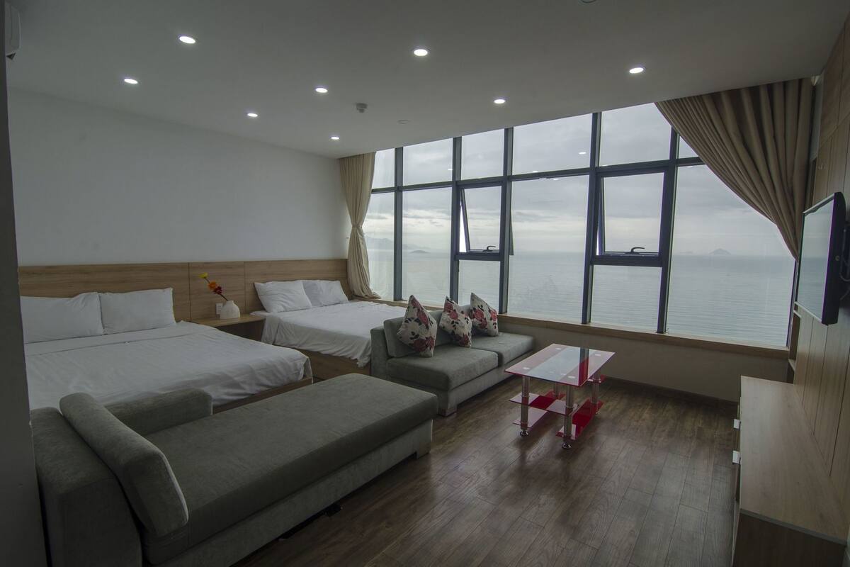 A hotel room with two beds, a sofa, a small table, and a large window with a view of the sea.