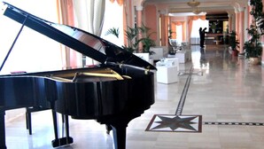Un piano de cola en un vestíbulo amplio con un diseño de piso en forma de estrella, mobiliario blanco y plantas en macetas.