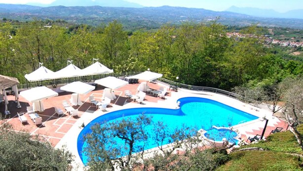 Una piscina con sillones de descanso y sombrillas, rodeada de árboles y con vistas a montañas distantes.