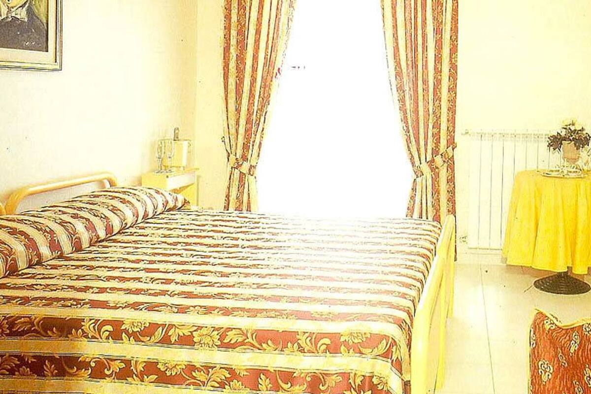 A room with a bed, a small table with a yellow tablecloth, and a painting on the wall.