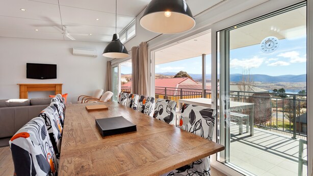 Uma área de refeições moderna com uma longa mesa de madeira, luminárias pendentes pretas e cadeiras estampadas. Uma televisão de tela plana está montada na parede, e é visível um balcão com vista para a paisagem através de grandes janelas.
