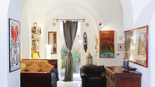 A cozy living room with a sofa, a leather armchair, and a wooden cabinet. There are framed artworks on the walls and a bookshelf in the background. - Caio Mario (Rome)