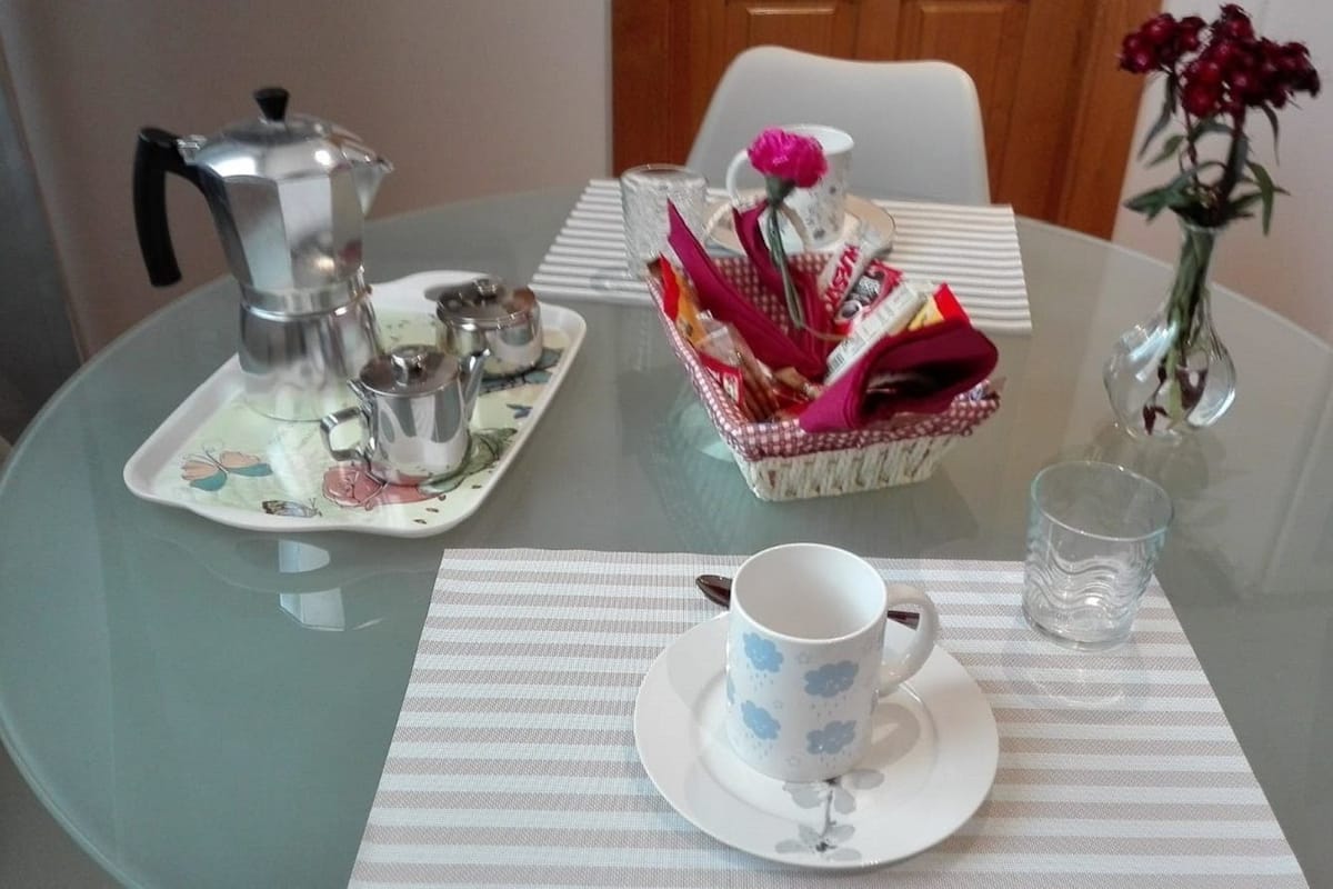 A round glass table with a white plate, a white cup, a glass of water, a teapot, and a basket of items.