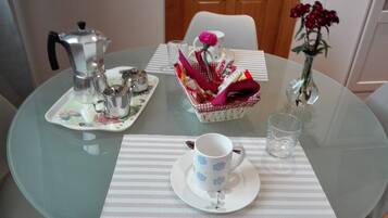 Una mesa redonda de vidrio con un plato blanco, una taza blanca, un vaso de agua, una tetera y una cesta de objetos.