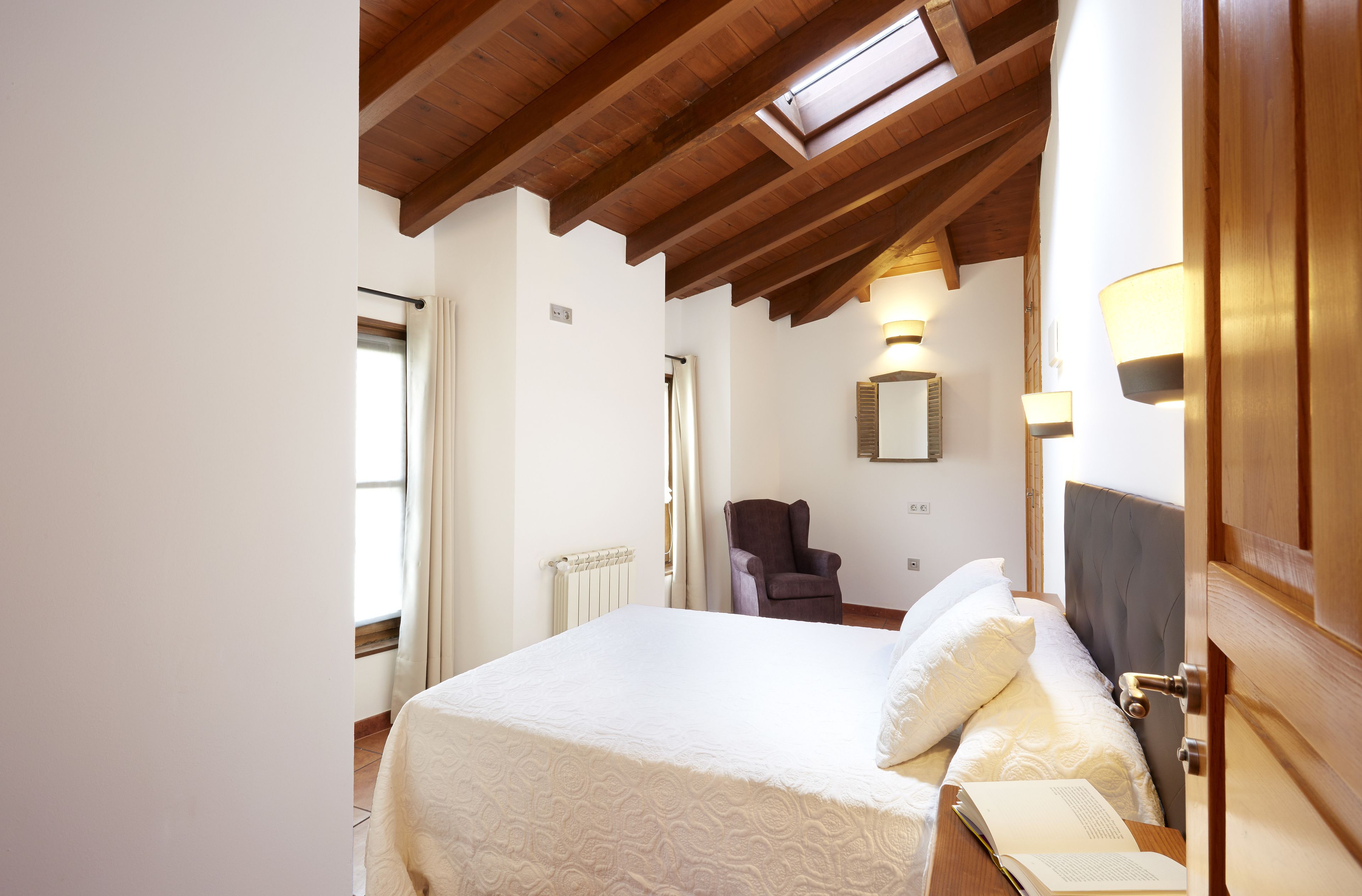 Un dormitorio con una cama grande, una silla, una mesita de noche con un libro, una ventana con cortinas y techo de madera.